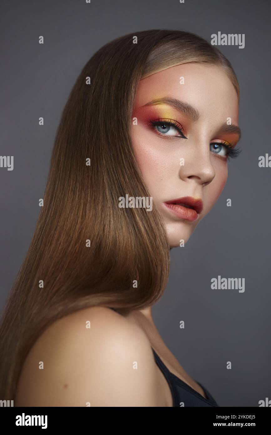 Auffällige Modellpositionen mit langem, glattem Haar und auffälligem, farbenfrohem Augen-Make-up in Rot- und Gelbtönen. Neutraler grauer Hintergrund betont das A Stockfoto