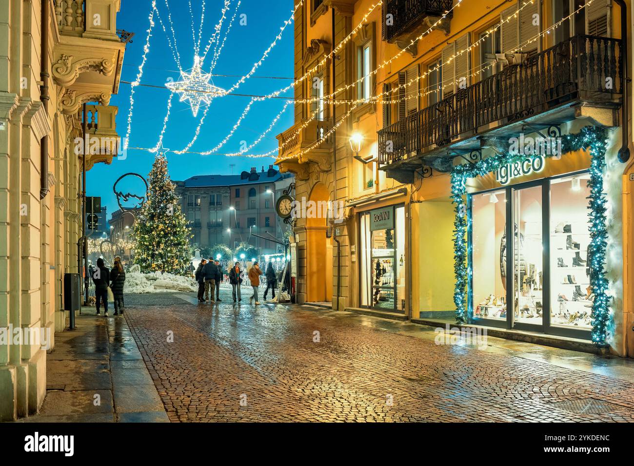 Die Menschen laufen entlang einer gepflasterten Straße, die mit festlichen Lichtern beleuchtet ist, wie ein Weihnachtsbaum im Hintergrund in Alba, Italien. Stockfoto