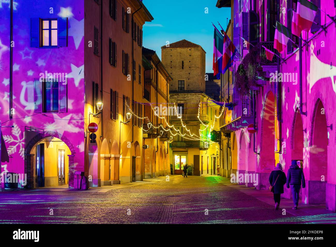 Kleine italienische Stadt Alba in der Abenddämmerung, mit schmaler Kopfsteinpflasterstraße und historischen Gebäuden, die abends von warmen Weihnachtslichtern beleuchtet werden. Stockfoto