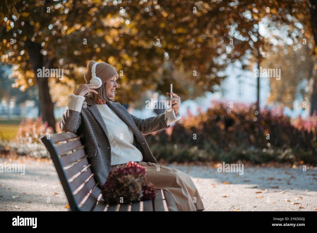 Porträt einer schönen Seniorin mit Kopfhörern im Herbstpark, Musik hören und ihr Telefon halten. Stockfoto