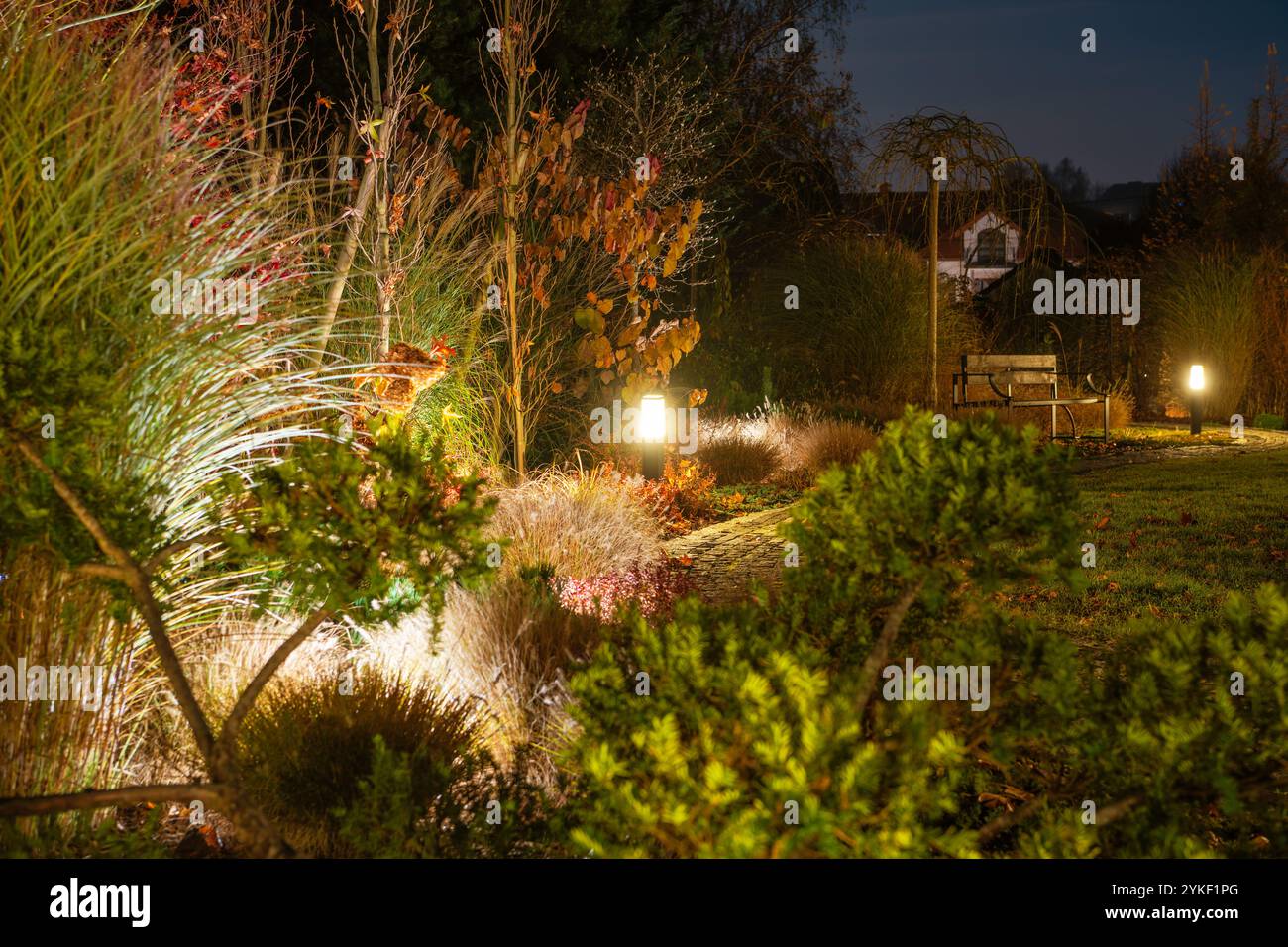 Ein Gartenweg wird von sanften Lichtern beleuchtet, umgeben von Herbstlaub und einer gemütlichen Bank unter dem Abendhimmel, die eine ruhige Atmosphäre schafft. Stockfoto