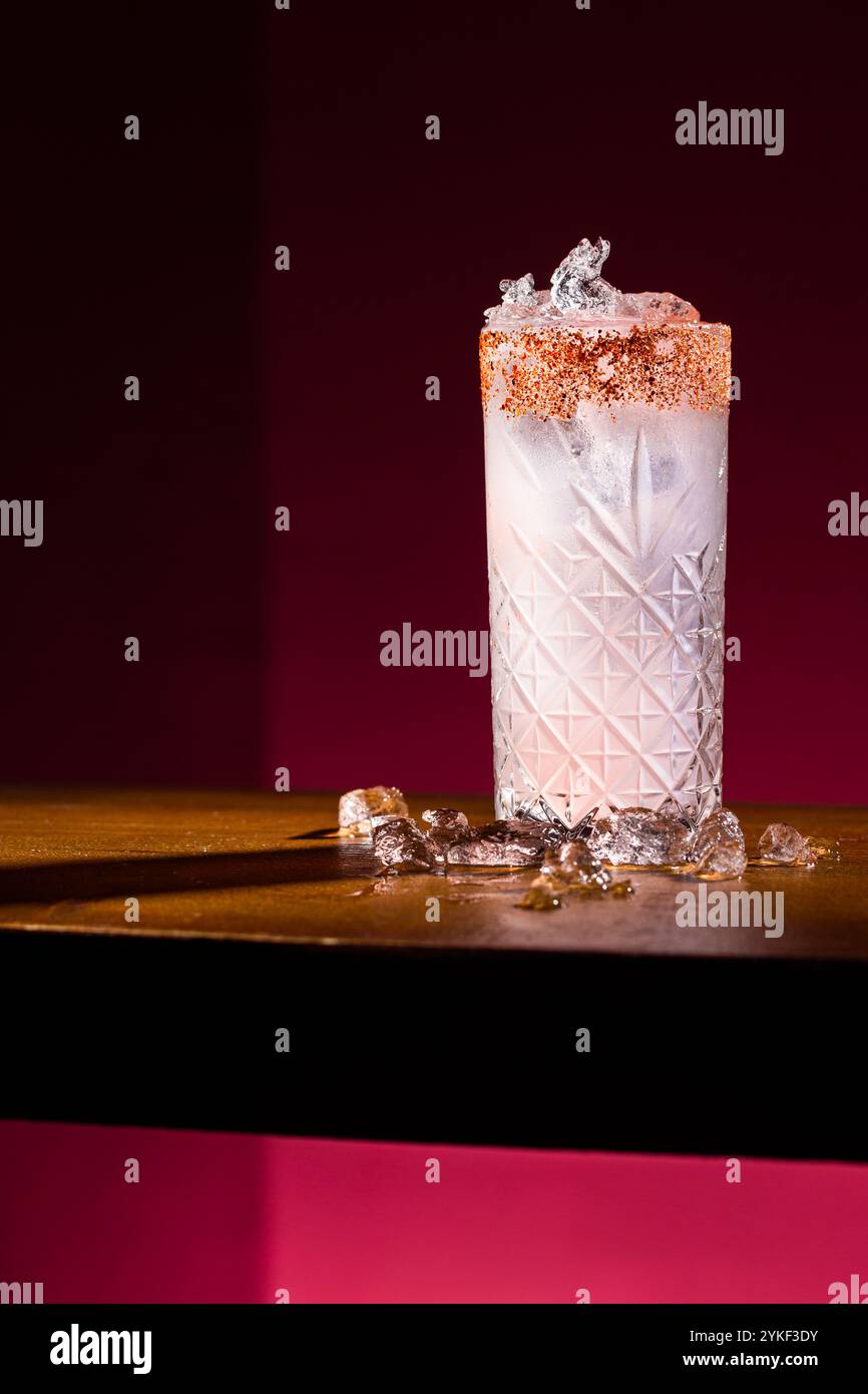 Ein atemberaubender Paloma-Cocktail mit gesalzenem Rand sitzt auf einer Holzoberfläche, umgeben von Eiswürfeln. Der leuchtend rosa Hintergrund verstärkt die erfrischende Toilette Stockfoto