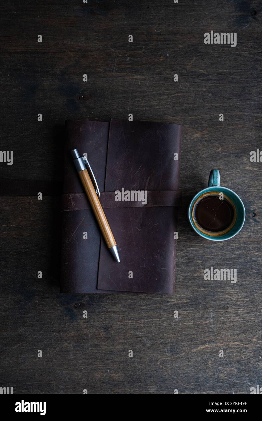 Blick von oben auf einen Holzschreibtisch mit einem ordentlich organisierten Ledernotizblock mit einem stilvollen Holzstift und einer vollen Tasse schwarzen Kaffees in der Nähe Stockfoto
