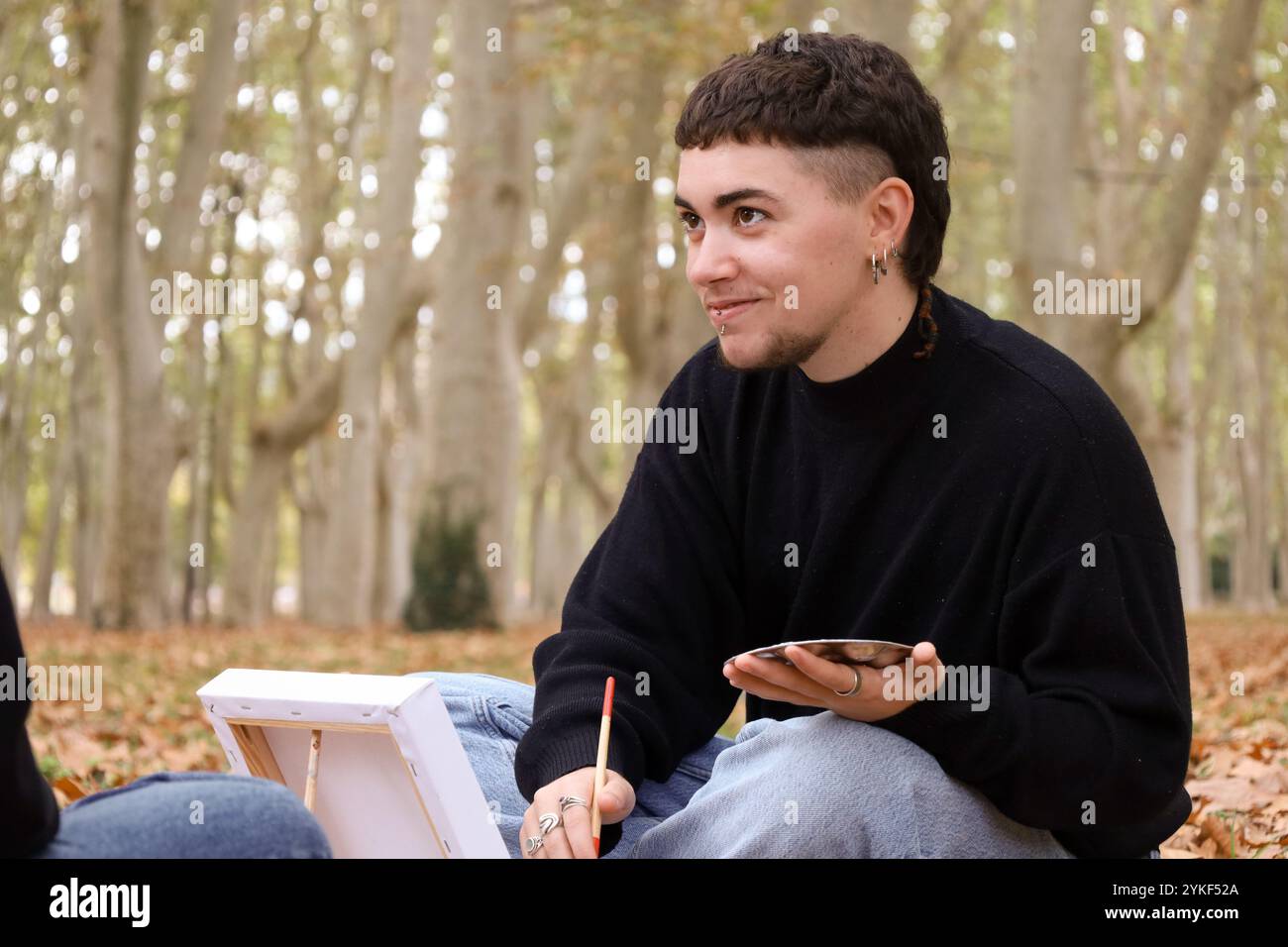Ein lächelnder Trans-Mann, der sich in einer kreativen Aktivität im Freien, umgeben von einem ruhigen Herbstwald, engagiert, symbolisiert Freude und LGBTQIA+ Stolz Stockfoto