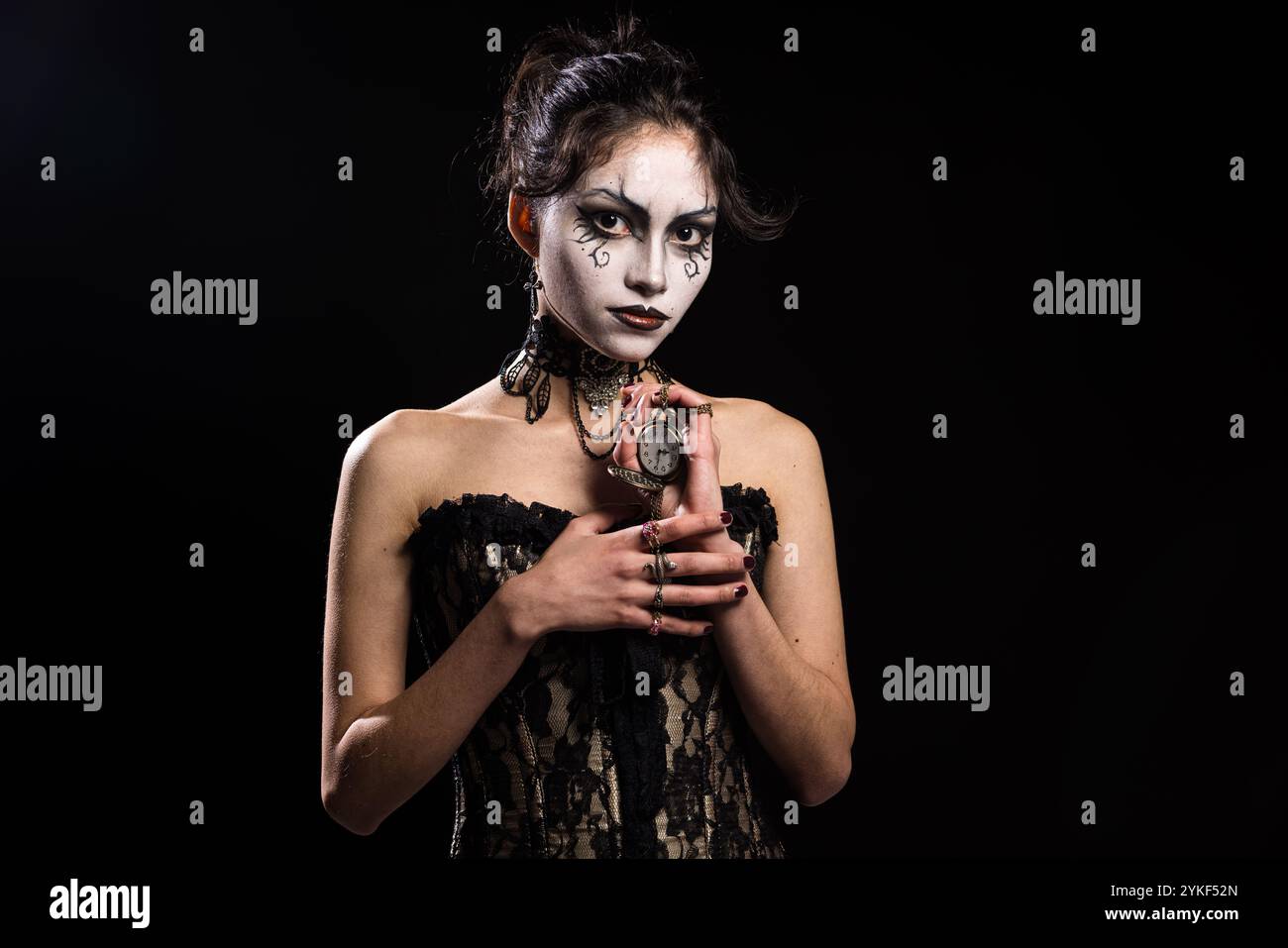 Eine gotische Frau in einem schwarzen Spitzenkorsett steht vor einem dunklen Hintergrund, ihr dramatisches Make-up, eleganter Schmuck und ihre Taschenuhr betonen einen myst Stockfoto