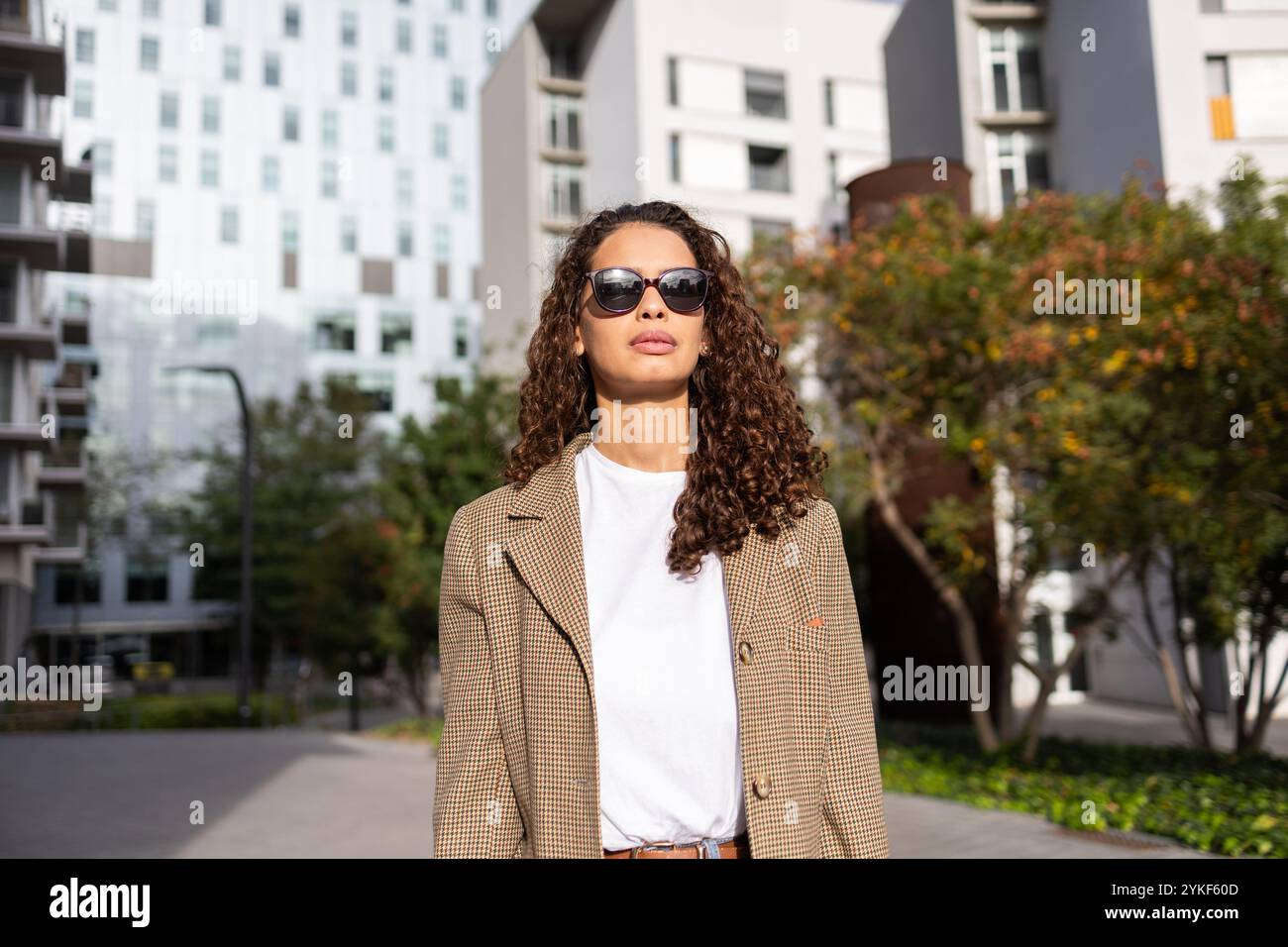 Eine elegante brasilianische Geschäftsfrau mit Sonnenbrille und Jacke steht selbstbewusst in einem urbanen Ambiente mit modernen Gebäuden im Hintergrund Stockfoto