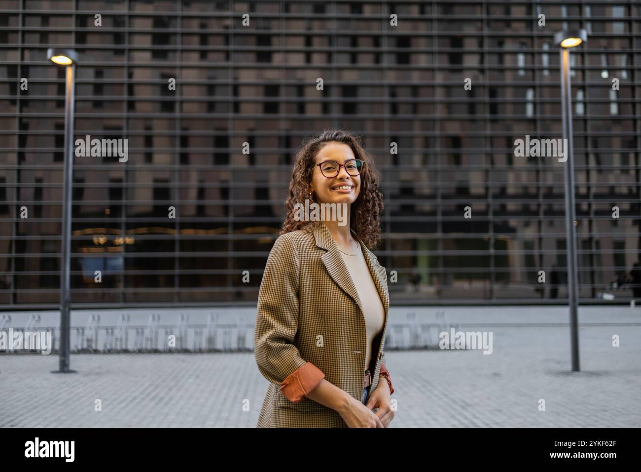 Eine junge brasilianische Geschäftsfrau mit lockigem Haar und Brille, die einen stylischen karierten Blazer trägt, lächelt in einer urbanen Umgebung mit modernem AR Stockfoto