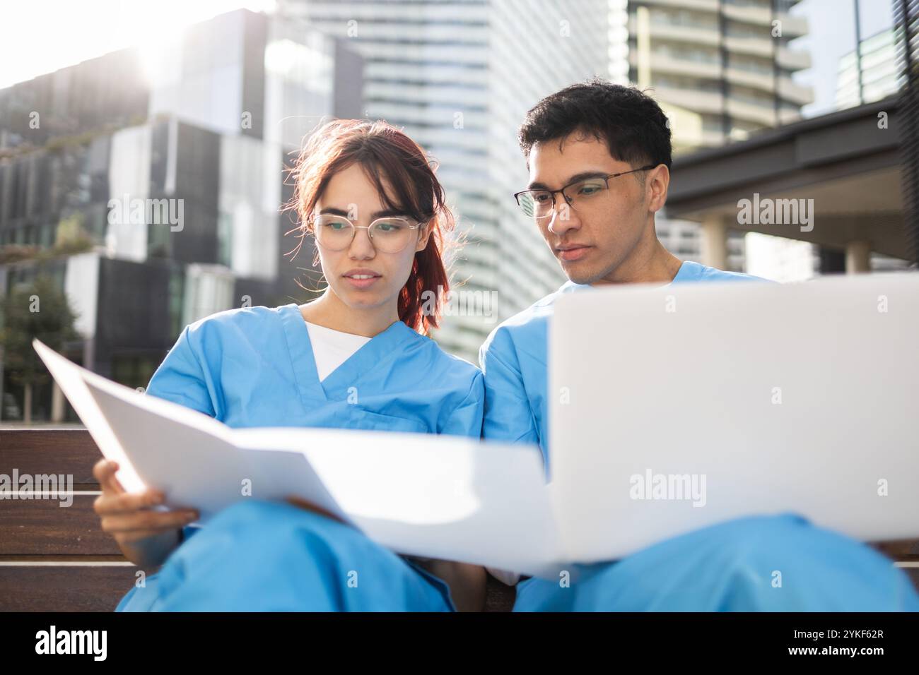 Zwei Schwesternstudenten in blauem Scrubs konzentrieren sich auf das Studium im Freien und konsultieren sowohl einen Laptop als auch Papierdokumente, vor einem modernen urbanen Hintergrund Stockfoto