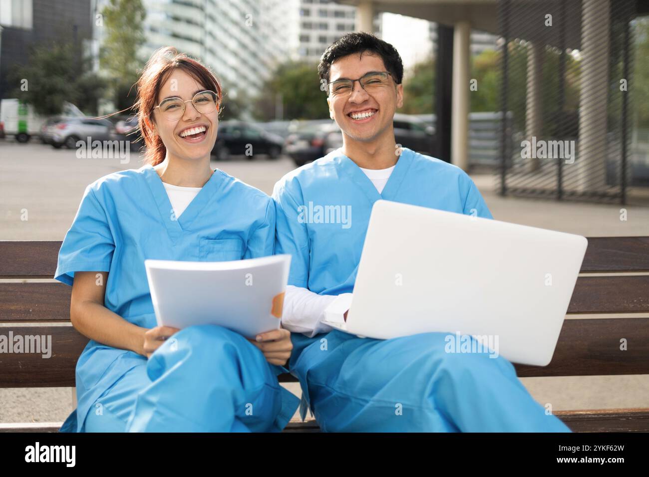 Zwei lächelnde Pflegestudenten, ein Mann und eine Frau, sitzen draußen auf einer Bank, studieren von Papieren und einem Laptop in ihren blauen Schutten Stockfoto