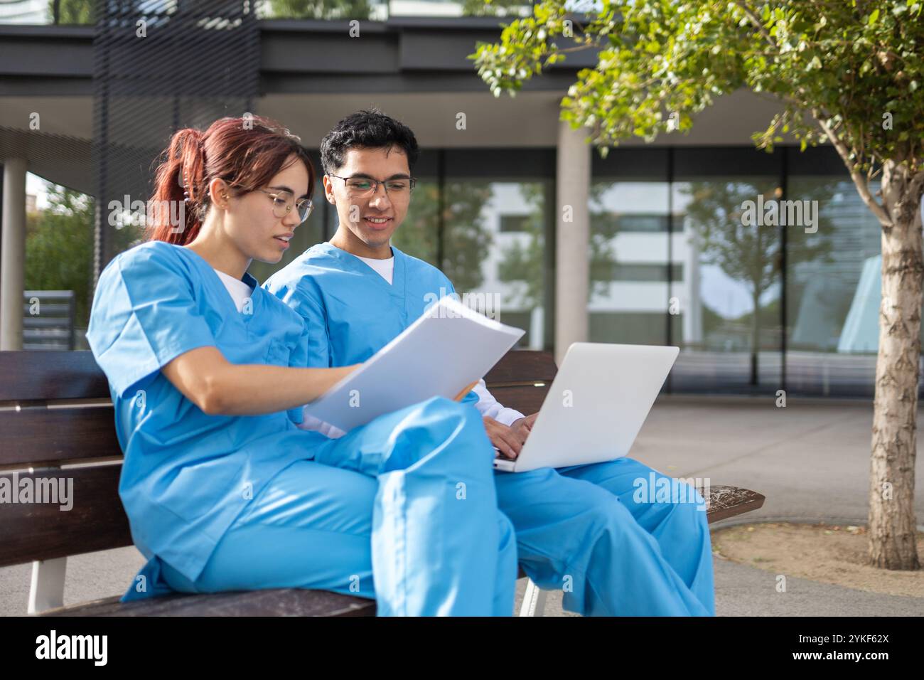 Zwei Schwesternschüler, ein Mann und eine Frau, in blauen Schutten sitzen vor einem Campus-Gebäude und arbeiten an ihren Aufgaben, diskutieren Notizen und benutzen einen Laptop f Stockfoto