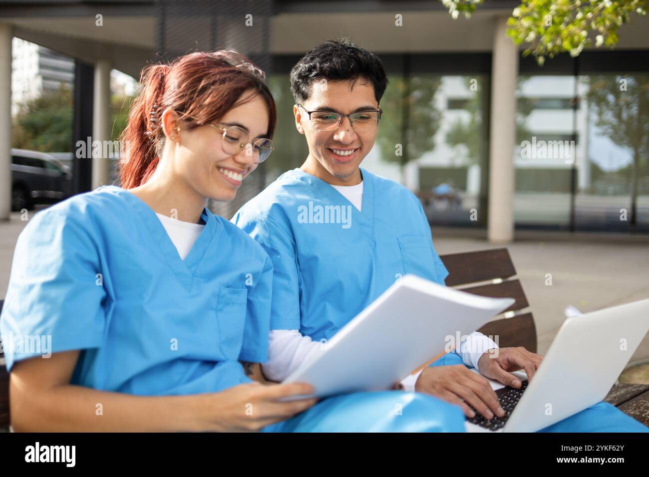 Zwei lächelnde Pflegestudenten, ein Mann und eine Frau, sitzen draußen auf einer Bank, studieren von Papieren und einem Laptop in ihren blauen Schutten Stockfoto