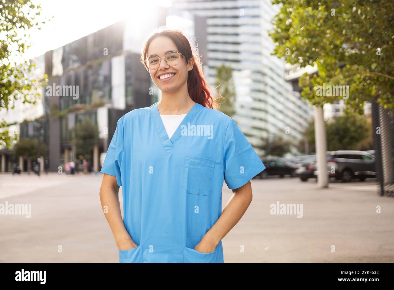 Eine fröhliche junge Frau in blauen Krankenschwester-Peelings steht selbstbewusst im Freien und verkörpert Optimismus und Professionalität in einem modernen urbanen Umfeld Stockfoto