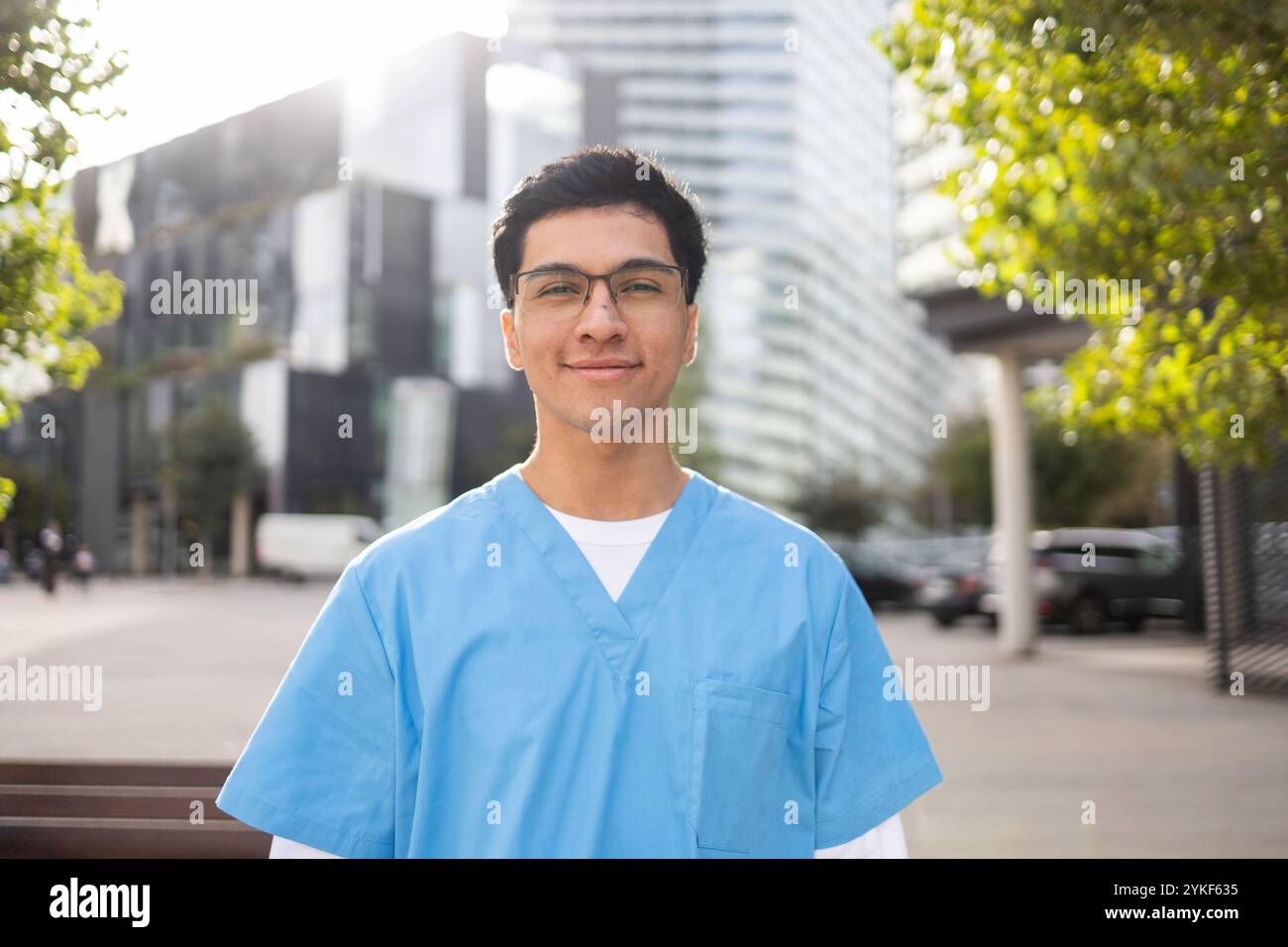 Ein junger Schwesternschüler in blauem Gewand lächelt selbstbewusst, steht draußen, mit einem verschwommenen Stadtbild Hintergrund Stockfoto