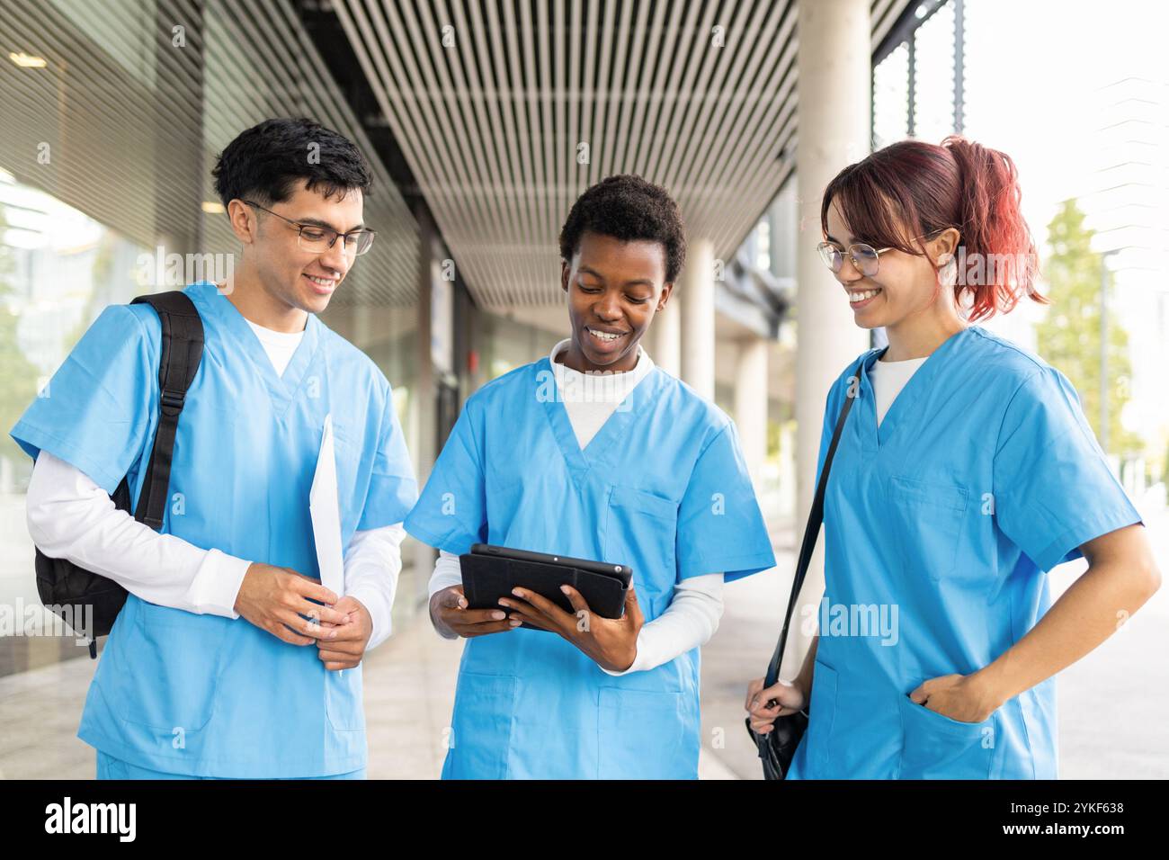 Eine vielfältige Gruppe von Pflegestudenten, zwei Frauen und ein Mann, führen sich mit einem Tablet vor einem modernen Gebäude, das in blauen Schuppen gekleidet ist, in eine lebhafte Diskussion Stockfoto