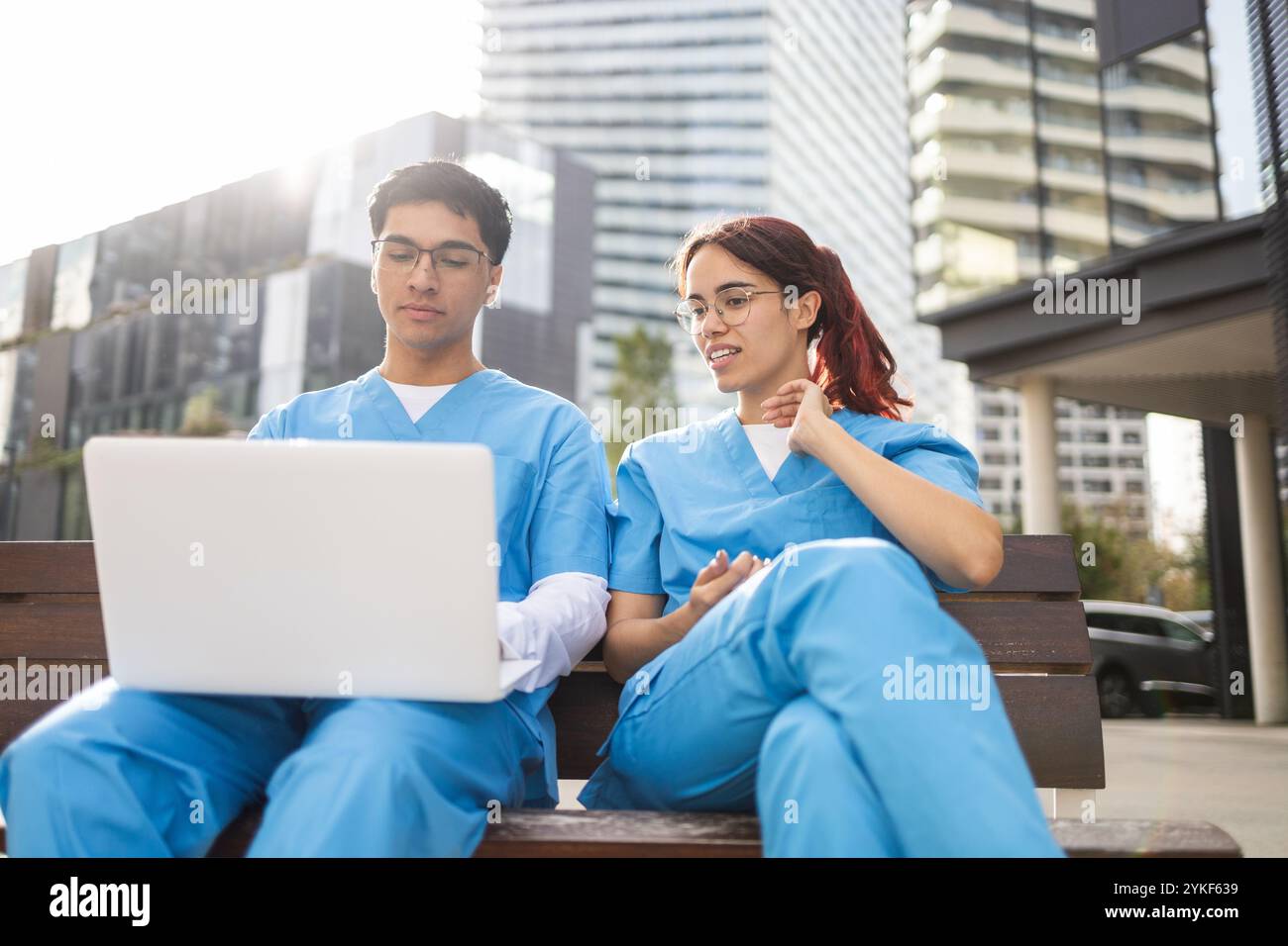 Zwei junge Krankenpflegeschülerinnen und -Studenten in blauen Schutten setzen sich mit einem Laptop auseinander, während sie draußen sitzen und akademische Inhalte in einem urbanen Umfeld diskutieren Stockfoto