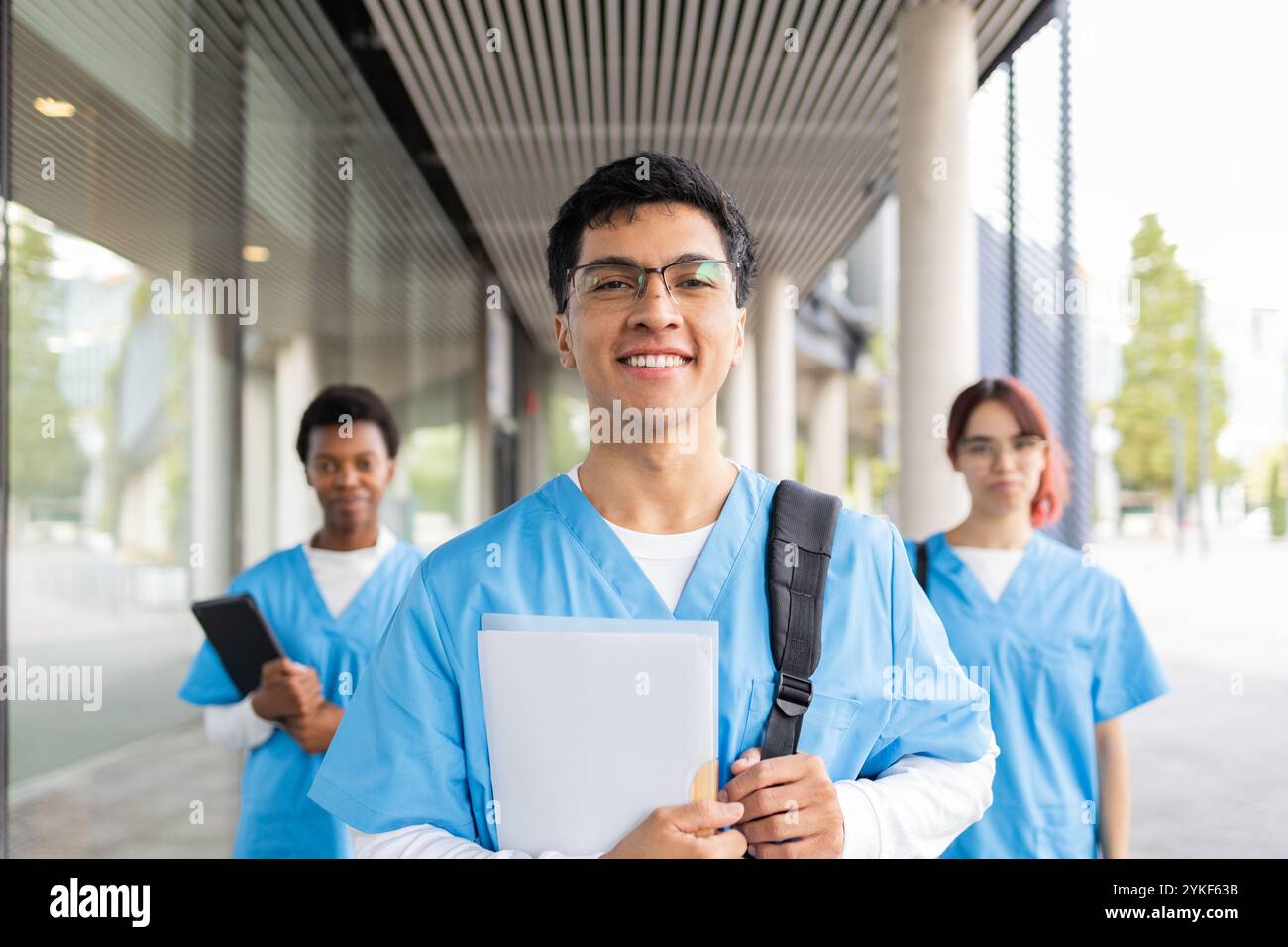 Fröhliche Pflegestudenten in blauen Schutten, mit Büchern und Taschen, selbstbewusst aus ihrer Medizinstudium gehen, bereit für ihre nächste Klasse Stockfoto