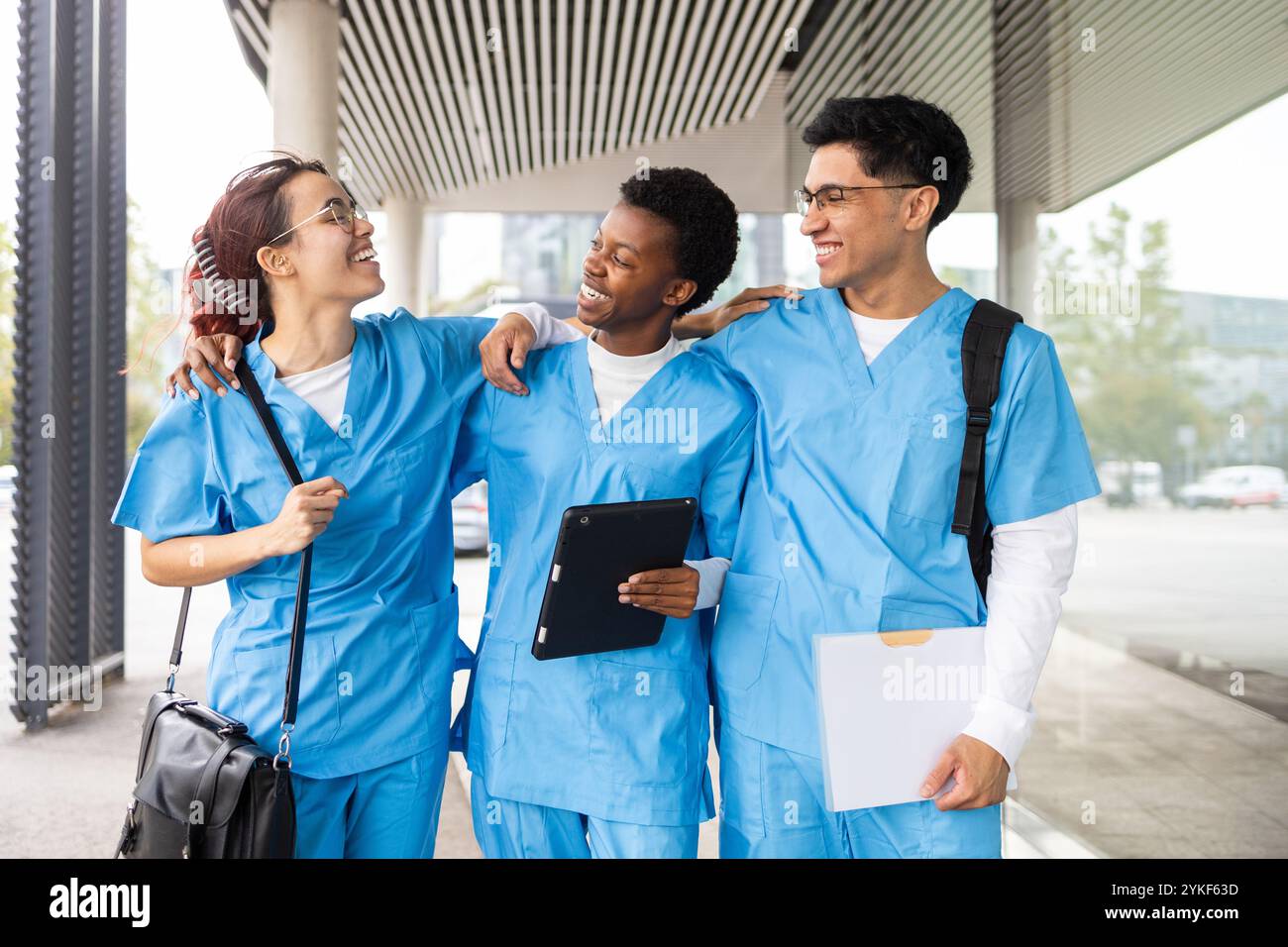 Drei multikulturelle Pflegestudenten in blauen Peelings interagieren gerne und teilen ihre Notizen außerhalb einer medizinischen Einrichtung Stockfoto