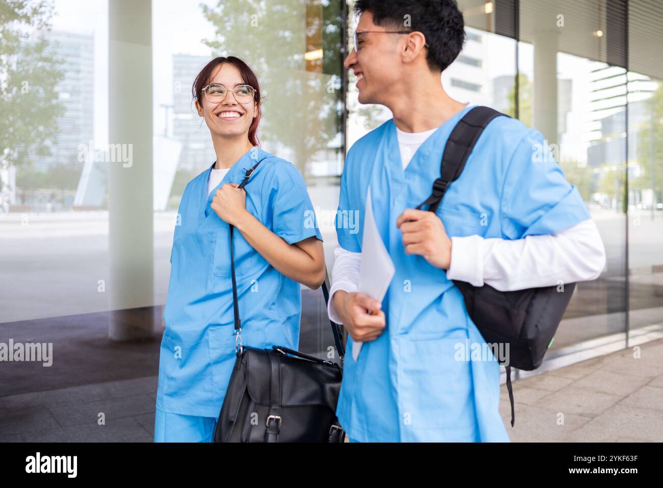 Zwei junge Schwesternstudenten, die sich vor einem modernen Universitätsgebäude in einer fröhlichen Unterhaltung unterhalten, beide tragen blaue Peelings und tragen Taschen mit einem C Stockfoto