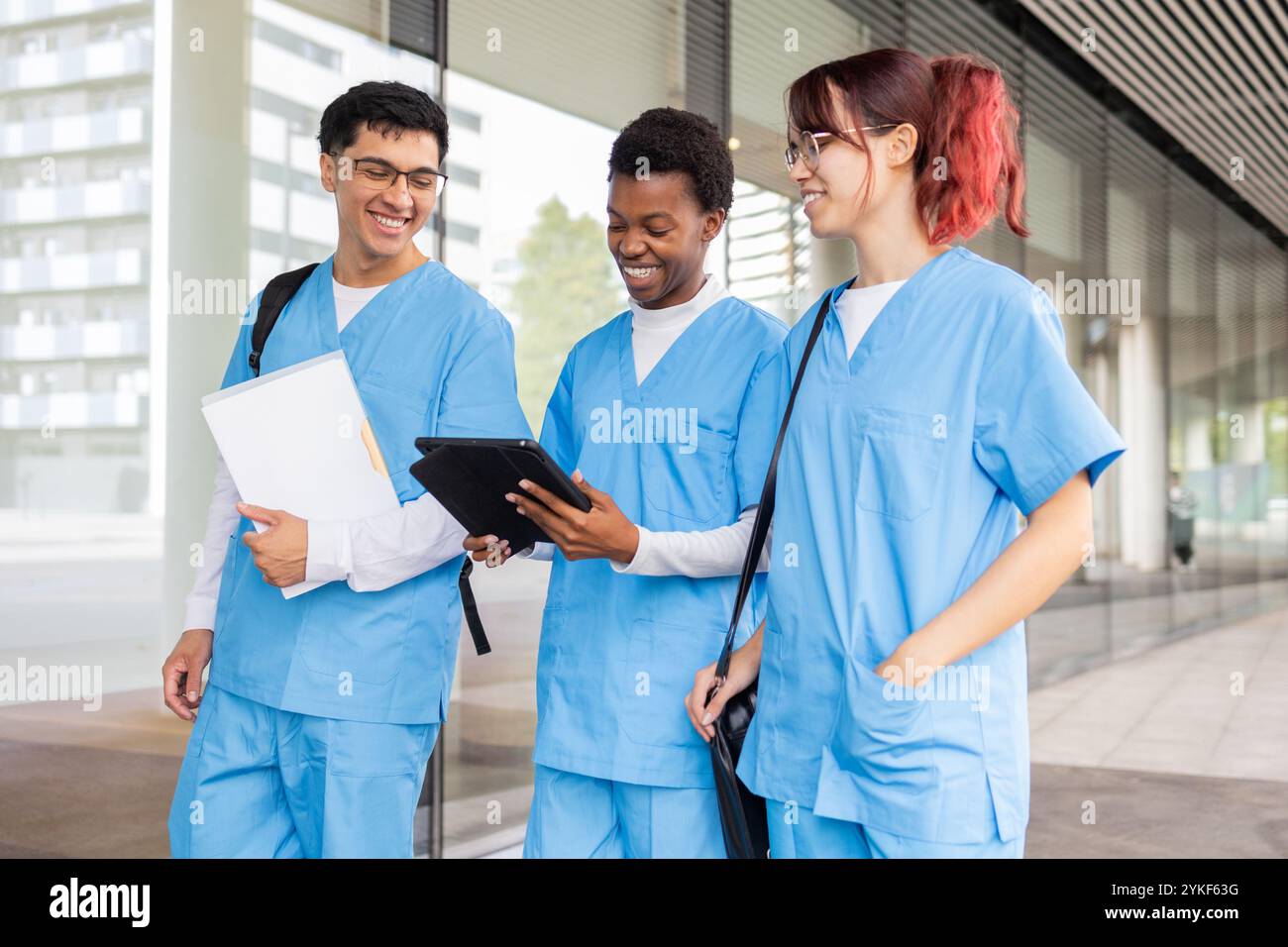 Drei Schwesternschüler in blauen Schutten teilen sich einen fröhlichen Moment und diskutieren mit einem digitalen Tablet außerhalb ihres medizinischen Campus über die Kursarbeit Stockfoto