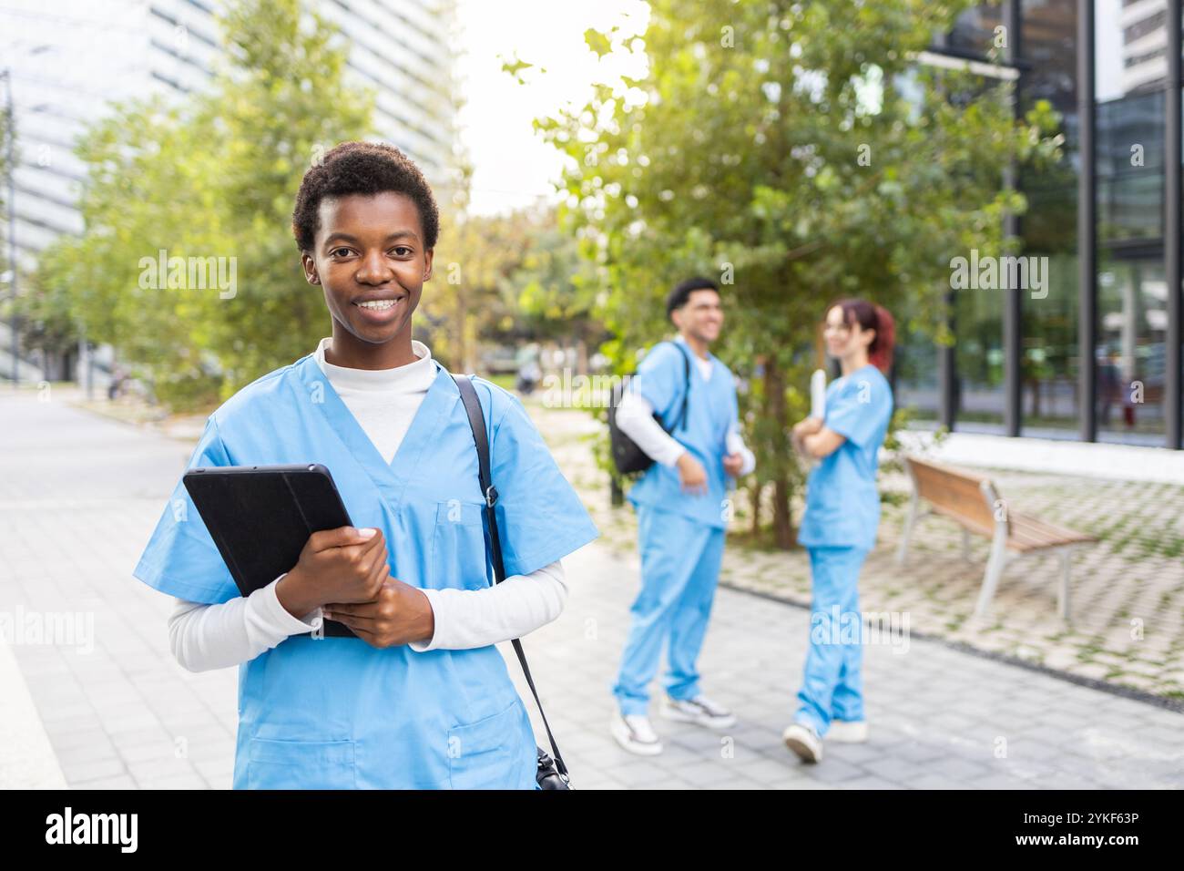 Eine junge Krankenschwesterstudentin in blauem Gewand lächelt mit einem Tablet, mit zwei Kollegen im Hintergrund, die sich auf einem lebhaften Universitätscampus unterhalten Stockfoto