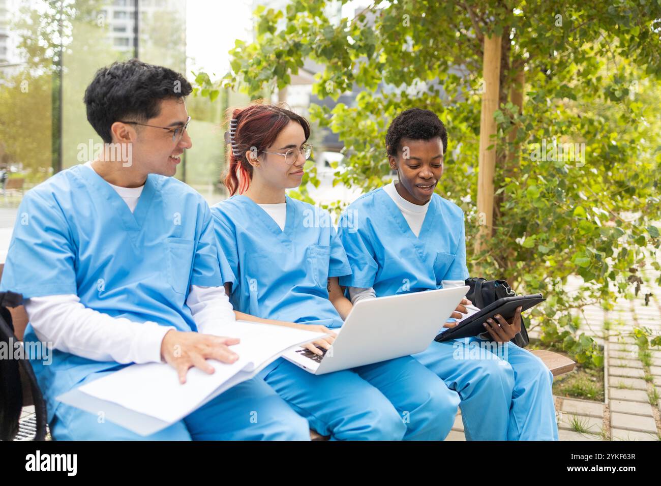 Drei Pflegeschülerinnen und Pfleger in blauen Schutten studieren gemeinsam mit einem Laptop und Notebooks in einem Park im Freien und zeigen eine Mutter Stockfoto