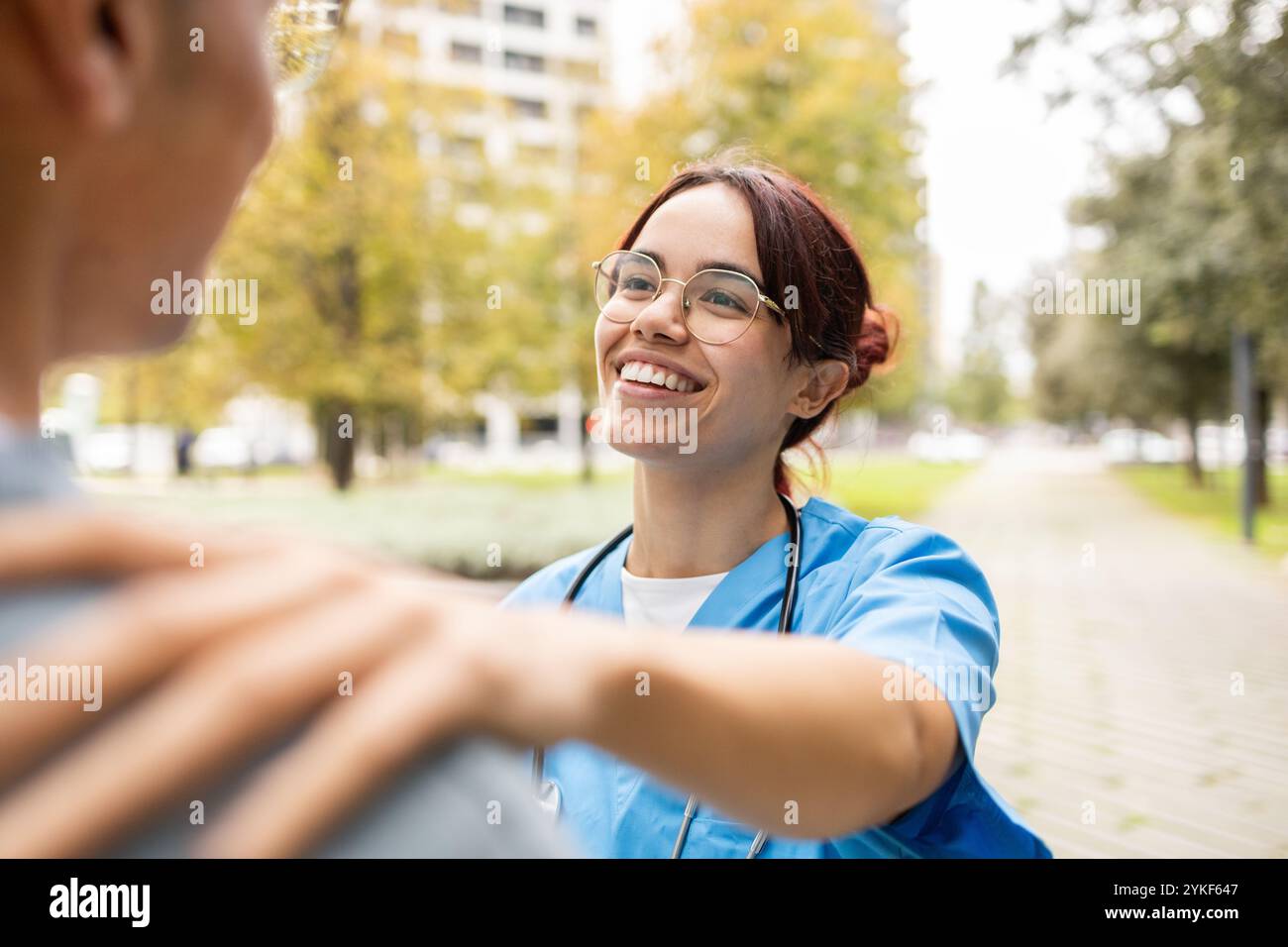 Eine fröhliche junge Krankenpflegeschülerin in blauen Schutten hilft einem Mann in einem Park, zeigt Mitgefühl und professionelles Engagement Stockfoto