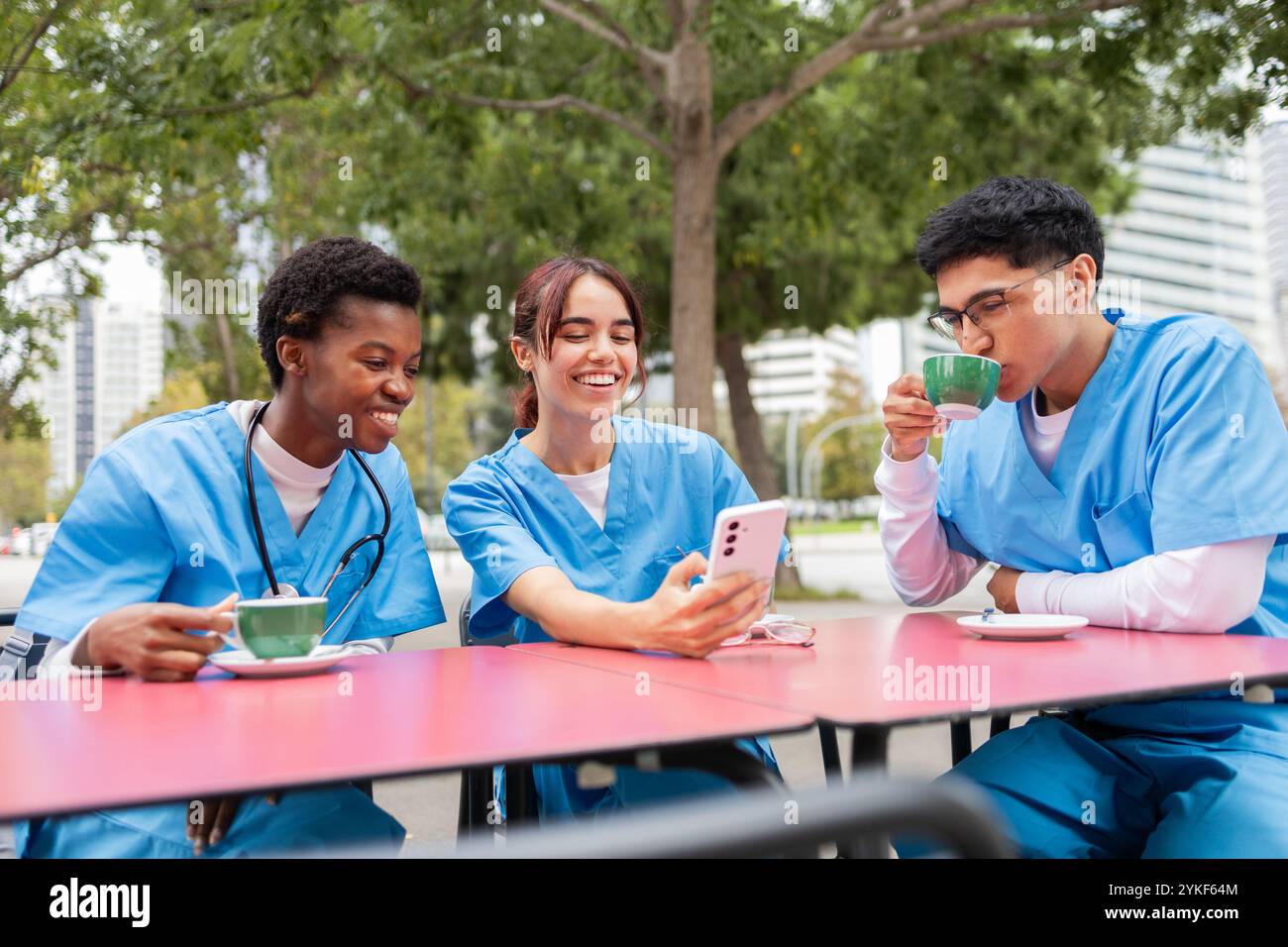 Eine Gruppe von drei Krankenpflegeschülern, die in blauen Peelings gekleidet sind, macht eine Pause im Freien einer lacht und hält ein Smartphone, ein anderer trinkt Kaffee, während der Stockfoto