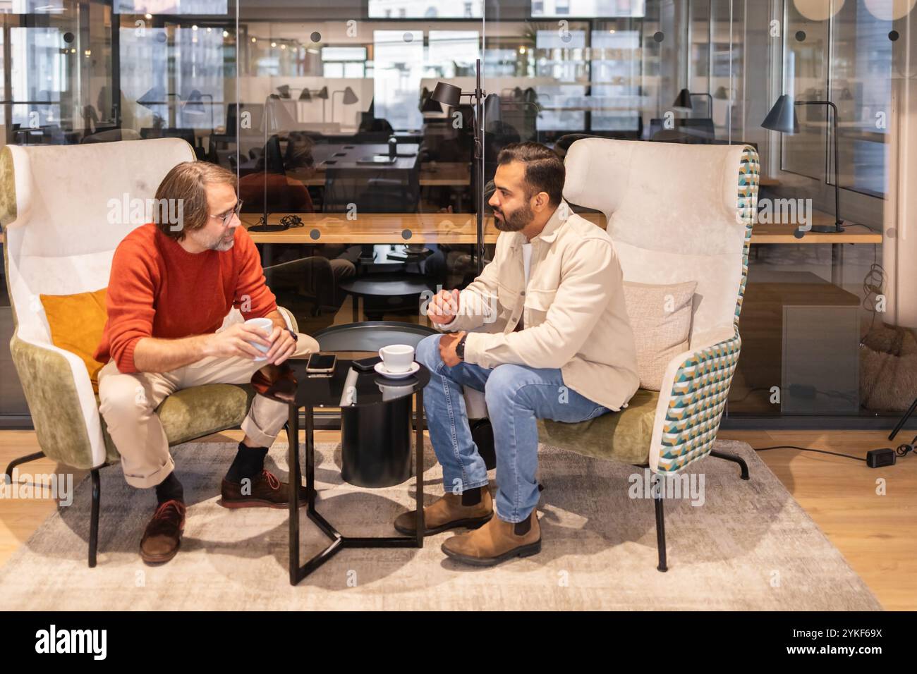 Zwei Kollegen unterhalten sich in einem stilvollen Coworking-Raum bei einem Kaffee in der entspannten Umgebung mit bequemen Stühlen und modernem Design Stockfoto