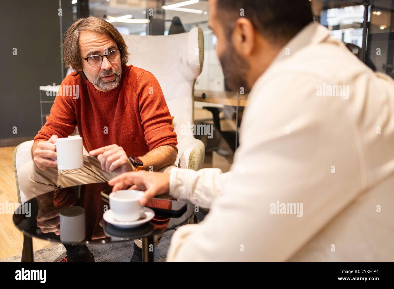 Zwei Kollegen führen in einem modernen Coworking-Raum ein entspanntes Gespräch bei einem Kaffee. Das Setting fördert die Zusammenarbeit mit Schwerpunkt auf Komfort Stockfoto