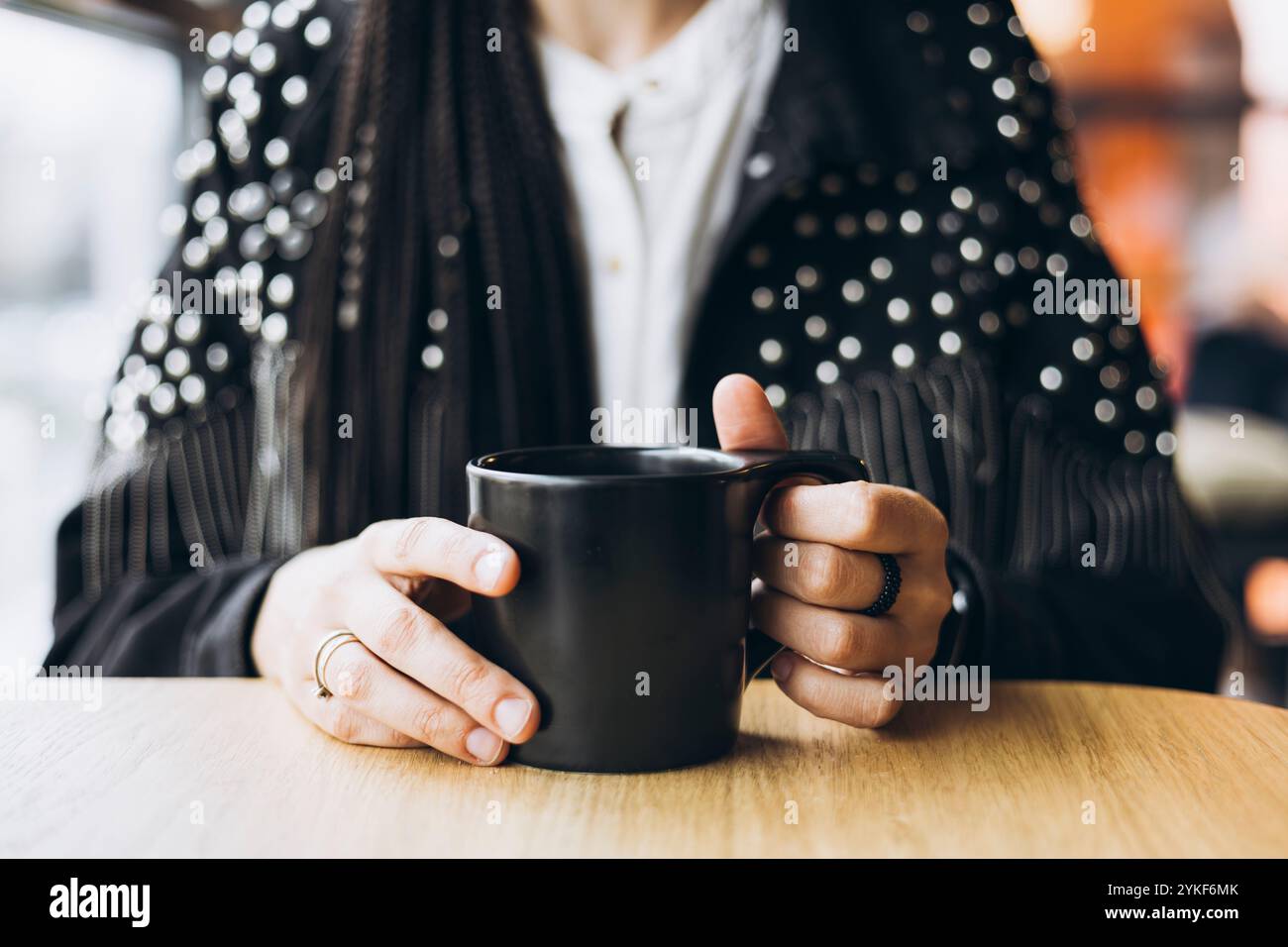 Eine stilvolle, nicht wiedererkennbare Frau mit Zöpfen schlürft in einem Café Kaffee aus einer schwarzen Tasse und zeigt ihre verzierte Jacke und ihre eleganten Ringe. Stockfoto