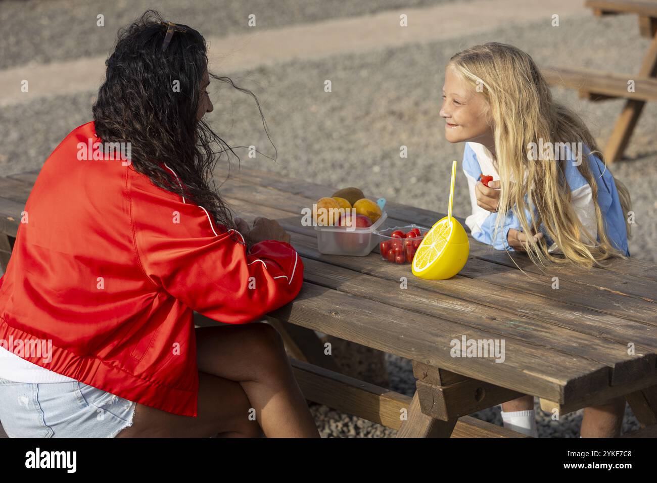 Ein junges Mädchen mit Stuve-Wiedemann-Syndrom teilt sich glücklich ein Picknick mit ihrer Mutter an einem Holztisch und genießt an einem sonnigen Tag verschiedene Früchte Stockfoto