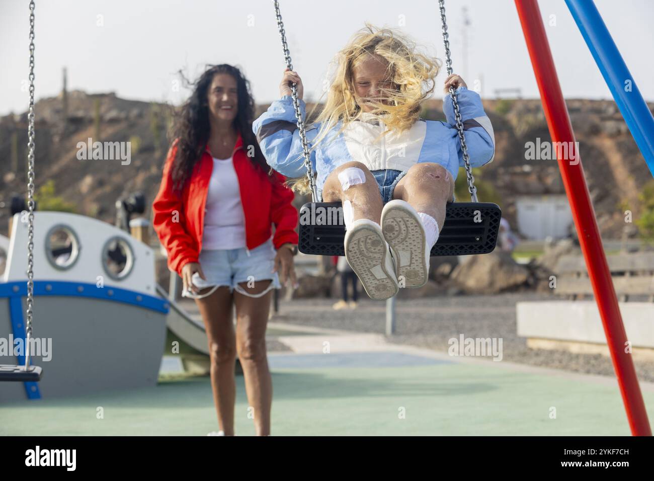 Ein fröhliches kleines Mädchen mit Stuve-Wiedemann-Syndrom schwingt unter der Unterstützung ihrer Mutter in einem sonnigen Park und spiegelt trotz eines Moments der Normalität wieder Stockfoto