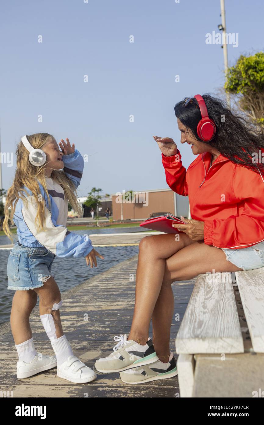 Eine junge Frau mit Stuve-Wiedemann-Syndrom teilt einen Moment der Freude mit ihrer Mutter, hört gemeinsam Musik entlang einer Promenade, während sie auf die Th blickt Stockfoto