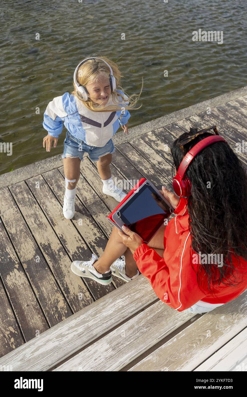 Eine glückliche junge Frau mit Stuve-wiedemann-Syndrom, lässig gekleidet und mit Kopfhörern, tanzt am Wasser, während ihre Mutter eine Tablette hält, hig Stockfoto