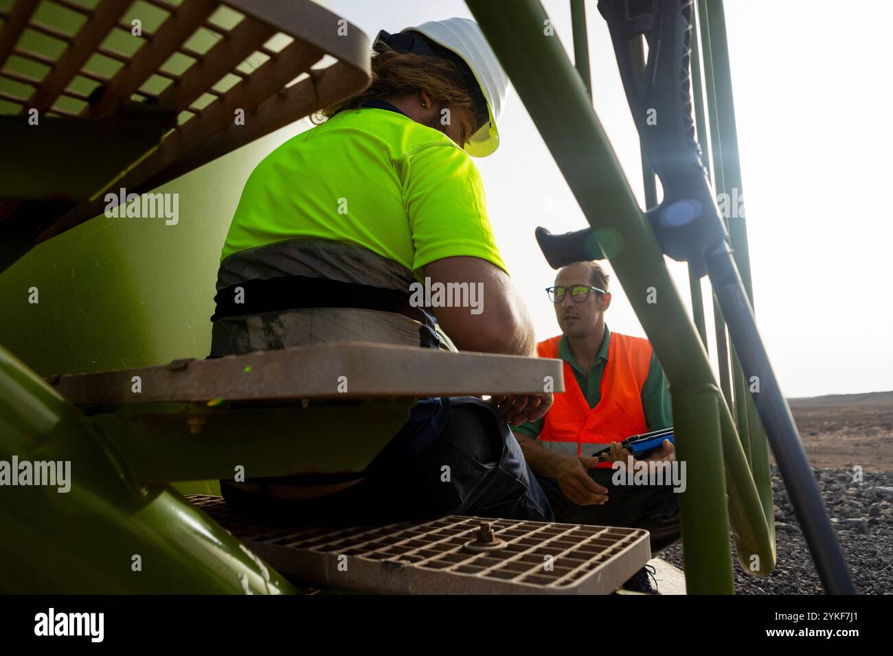 Zwei Arbeiter, die Helme und Warnwesten tragen, werden an der Basis einer Windkraftanlage in einem Windpark gesehen, in dem diskutiert wird, demonstrieren sie Stockfoto