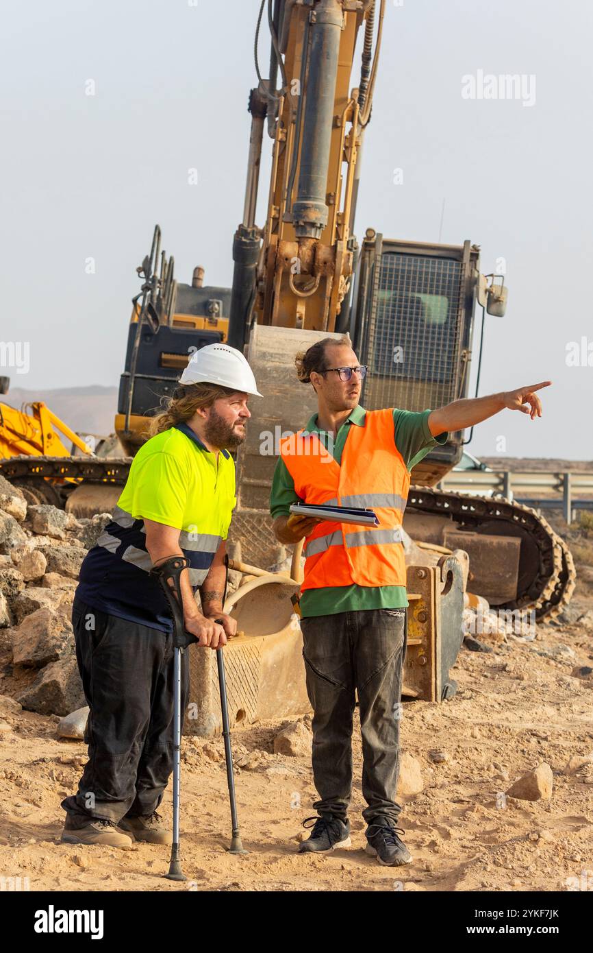 Ein behinderter Inspektor mit Krücken arbeitet mit einem Kollegen in einer Sicherheitsweste zusammen und zeigt Details auf einer Baustelle für erneuerbare Energien auf, empha Stockfoto