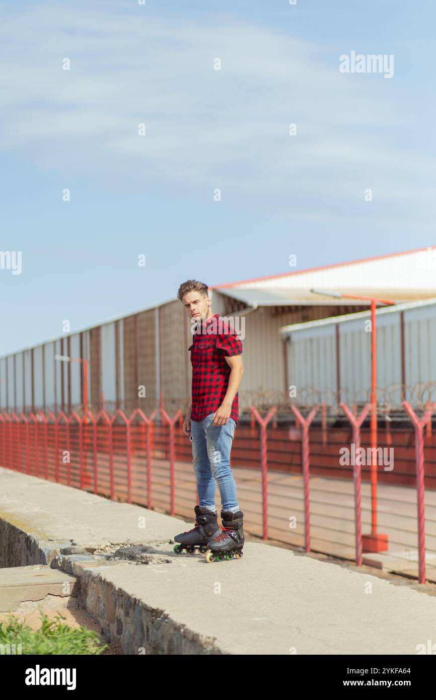 Ein junger Mann in einem roten karierten Hemd und Jeans steht selbstbewusst auf Inline-Skates und blickt von der Kamera weg, vor einem industriellen Hintergrund mit Stockfoto