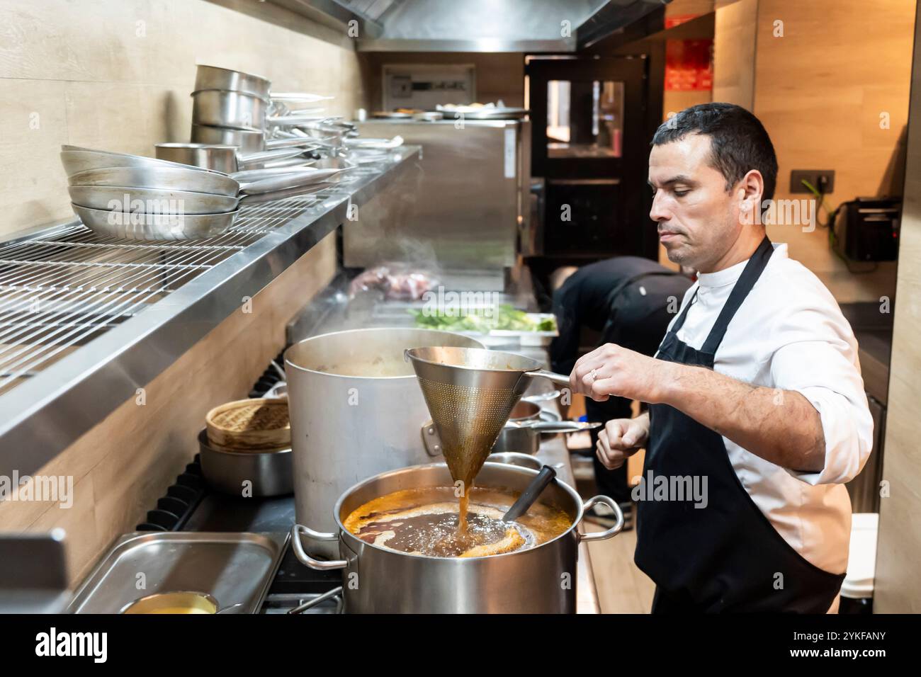 Der professionelle Koch hat sich darauf konzentriert, einen großen Topf Suppe in einer gut ausgestatteten Restaurantküche zu würzen, umgeben von verschiedenen Kochutensilien und App Stockfoto