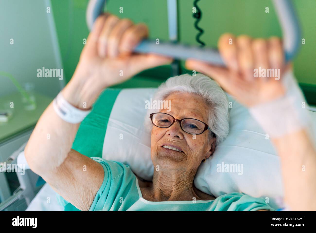 Eine ältere Frau, die in einem Krankenhausbett liegt und sich an einer Trapezstange festhält. Die Umgebung ist hell und sauber und spiegelt eine Gesundheitsumgebung wider Stockfoto