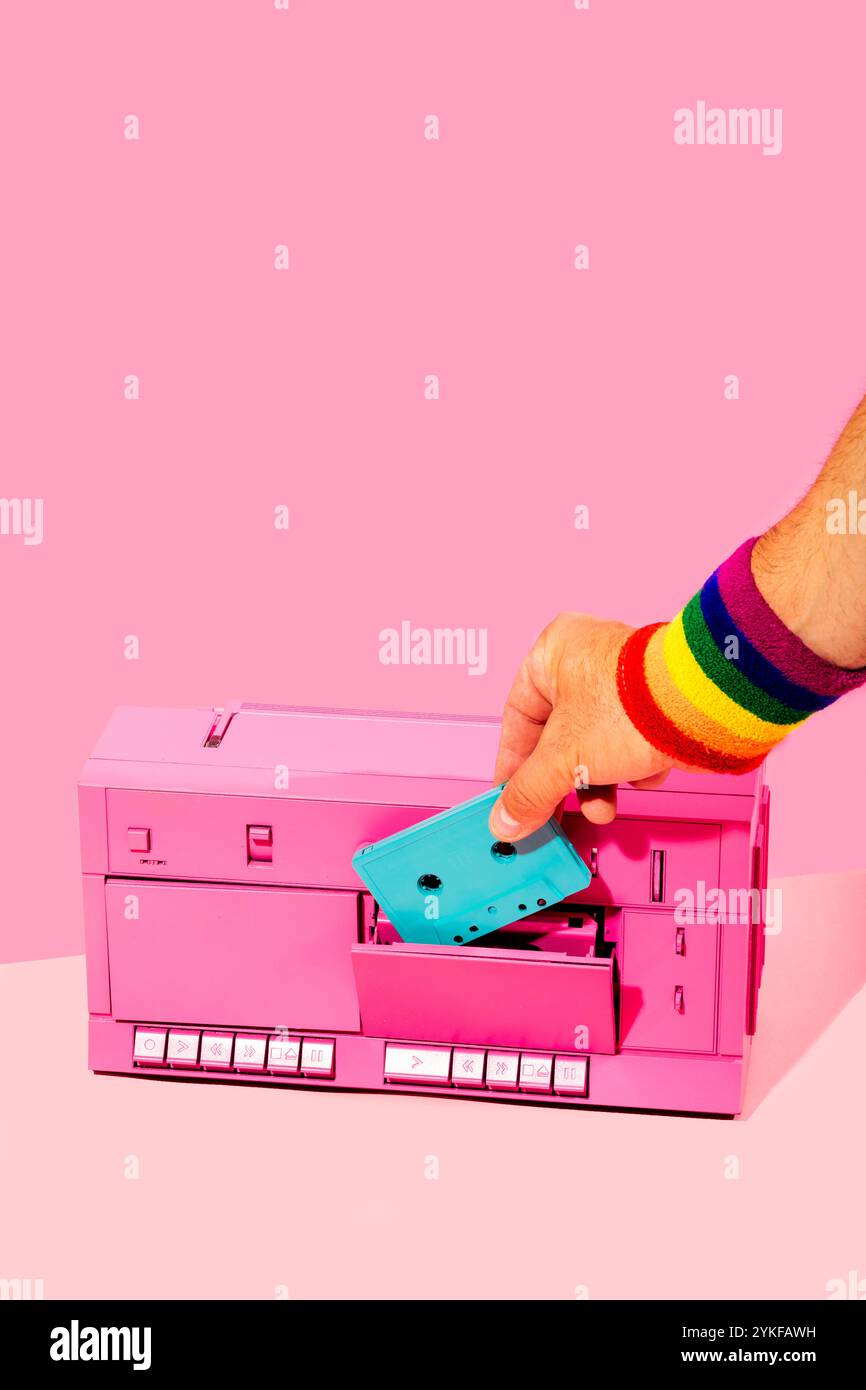 Eine Hand mit einem Regenbogenarmband fügt eine blaue Kassette in einen leuchtend rosa Retro-Kassettenspieler ein. Das Bild kombiniert ein nostalgisches Thema mit modernem LGBTQIA Stockfoto
