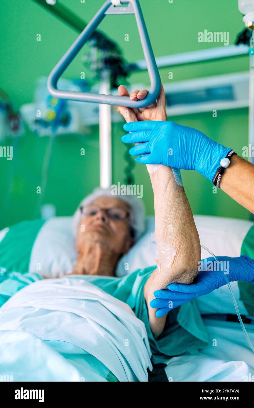 Eine ältere Frau liegt in einem Krankenhausbett, wobei ihr Arm von einem medizinischen Fachpersonal mit Schutzhandschuhen sanft gestützt wird Stockfoto