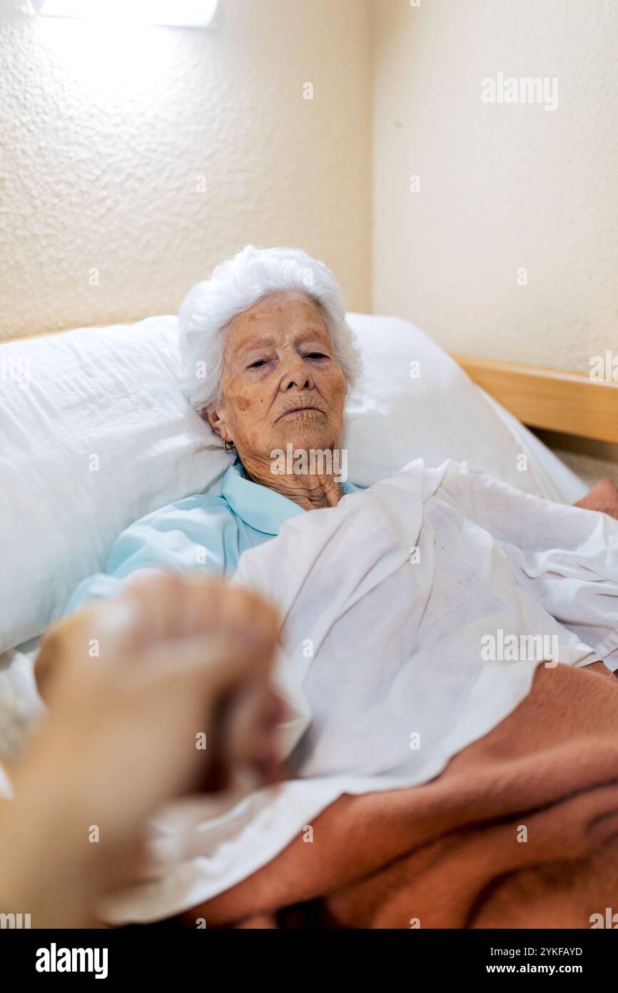 Eine ältere Frau liegt im Bett und hält die Hände mit jemandem außerhalb des Rahmens. Die Szene vermittelt ein Gefühl von Sorgfalt und Gesellschaft und hebt Themen von älteren Menschen hervor Stockfoto