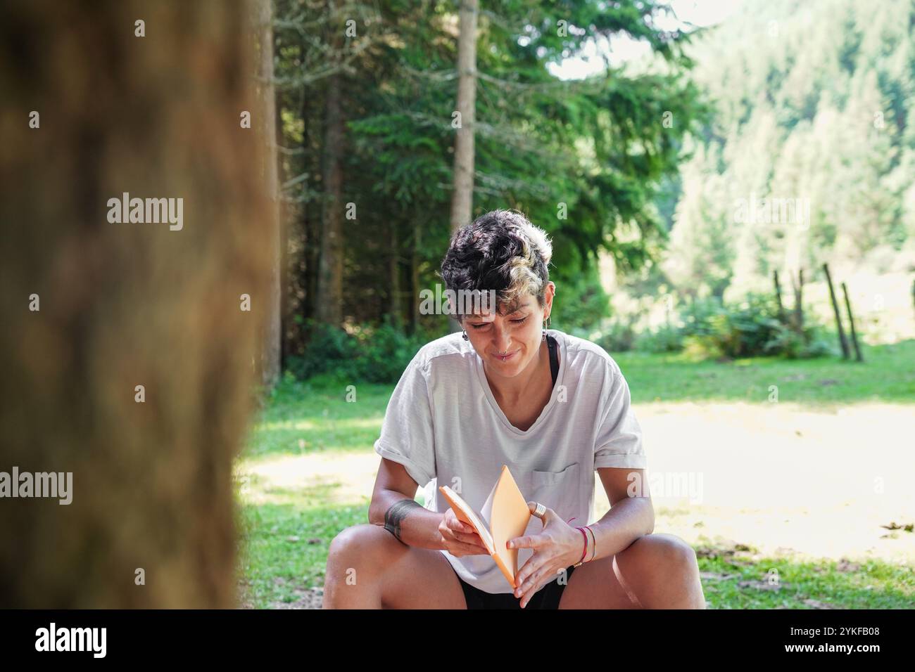 Ein nicht-binäres Individuum, das in das Lesen eines Buches vertieft ist, lässig in einem üppigen Waldgrund sitzt und ein Gefühl von Frieden und Ruhe vermittelt. Stockfoto