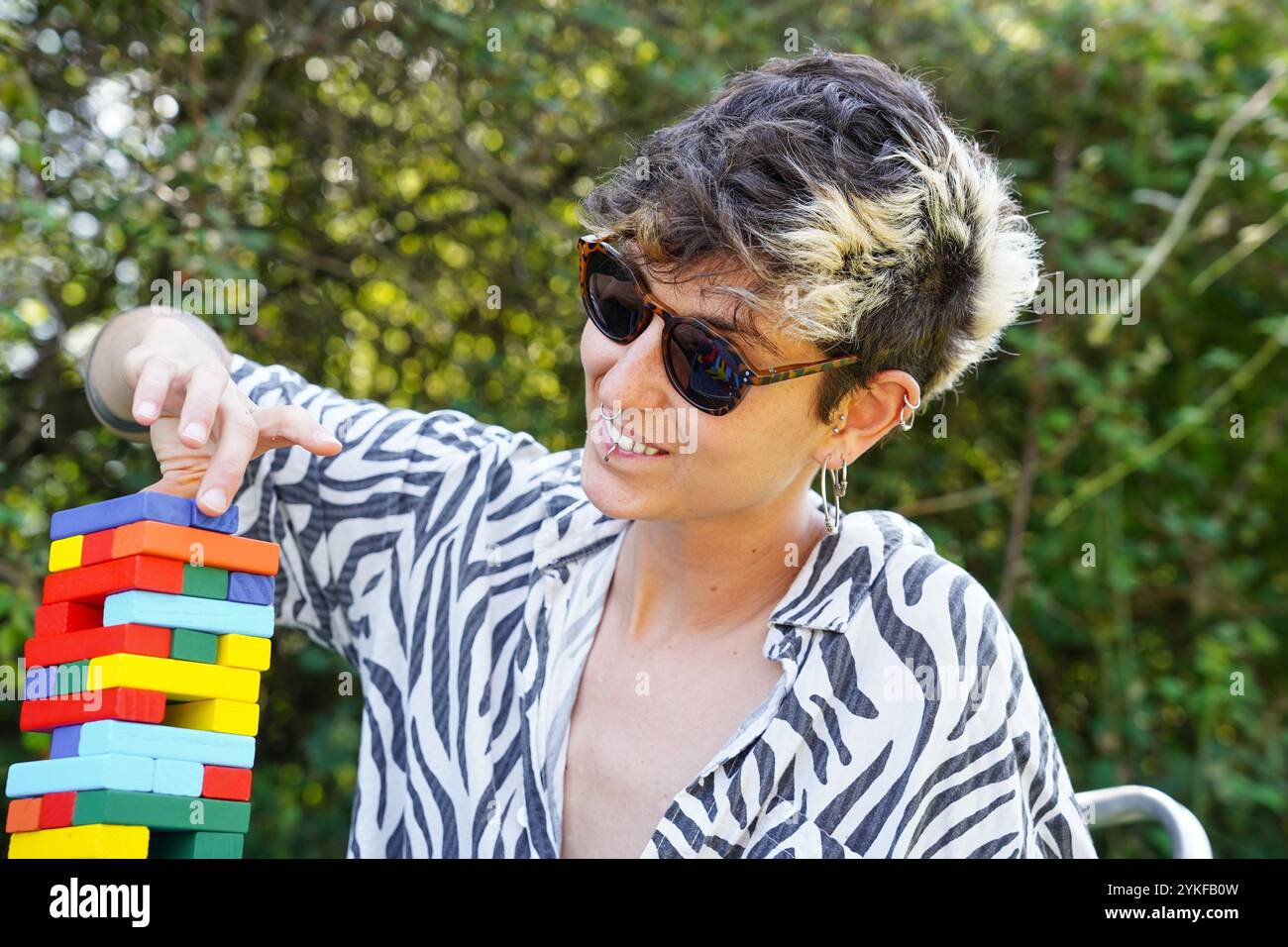 Ein nicht-binäres Individuum mit kurzen Haaren und Sonnenbrille spielt fröhlich mit einem lebendigen, mehrfarbigen Turm aus Blöcken im Freien, umgeben von G Stockfoto