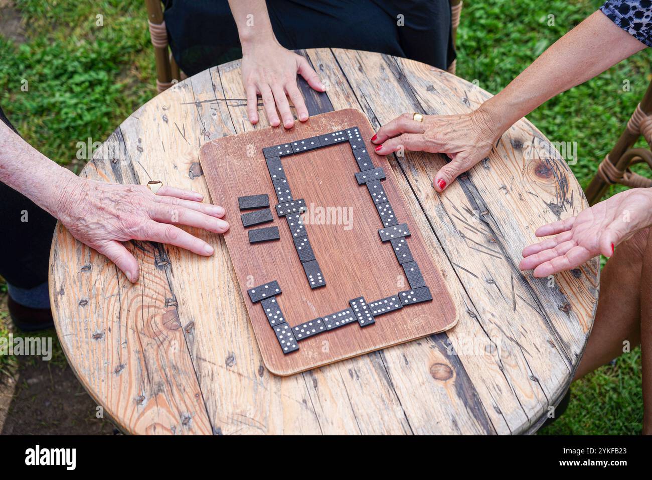 Eine abwechslungsreiche Familiengruppe, darunter Senioren und Jugendliche, spielt begeistert an einem Brettspiel um einen rustikalen Tisch im Freien. Ihre Hände, Stockfoto