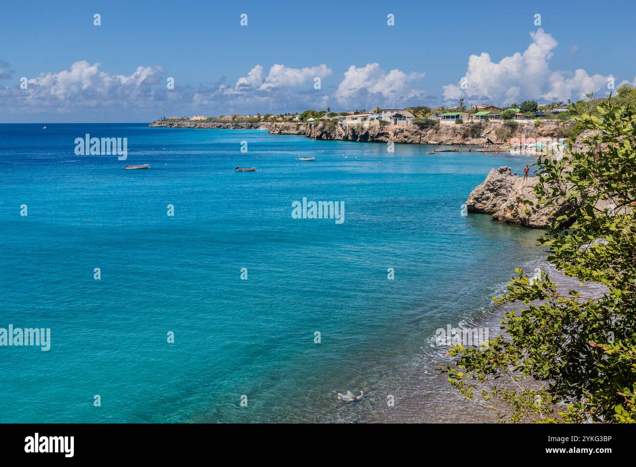 Die Bucht von Westpunt liegt im äußersten Nordwesten von Curaçao, ihre türkisfarbenen karibischen Gewässer sind mit Fischerbooten gesprenkelt, die vor Anker liegen. Auf dem Felsvorsprung rechts bereitet sich eine Person darauf vor, ins Meer zu springen. Die Klippen um Playa Forti in Westpunt sind bei Klippenspringern bekannt. Stockfoto