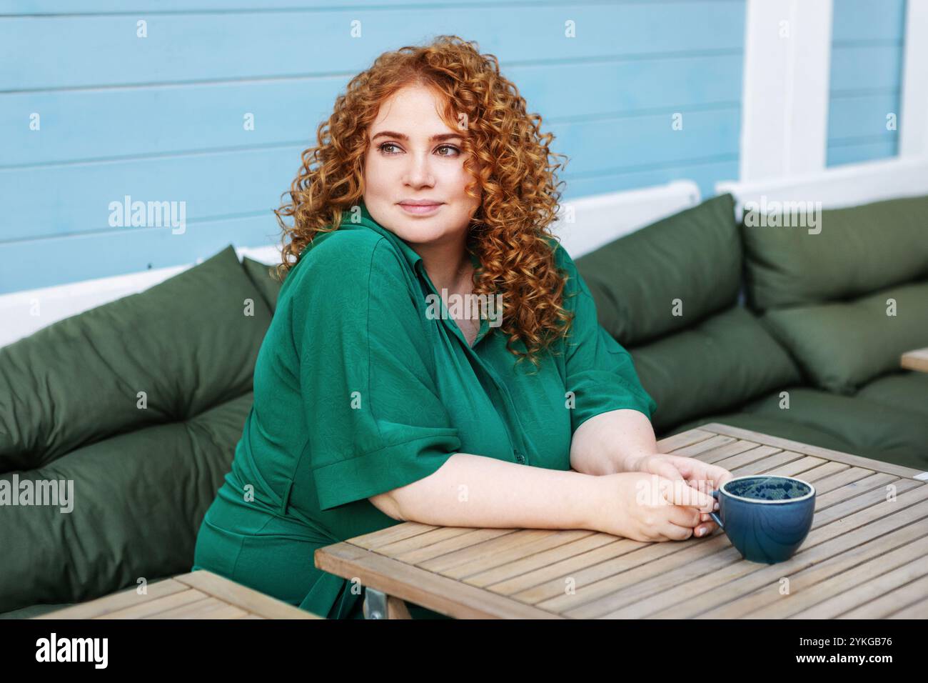 Schöne große Frau mit rotem Haar, die eine Tasse Kaffee genießt. Kurviges weibliches Model sitzt am Cafe-Tisch in grünem Kleid gegen helles buntes blaues W Stockfoto