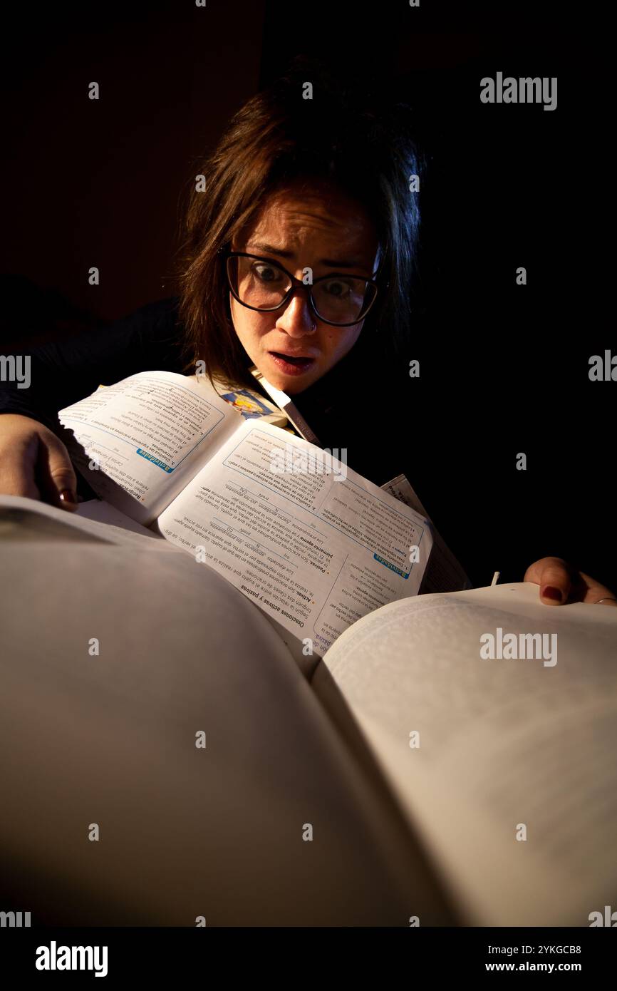 Gestresste junge Frau mit Brille studiert für Prüfungen, umgeben von Büchern in der Nacht Stockfoto