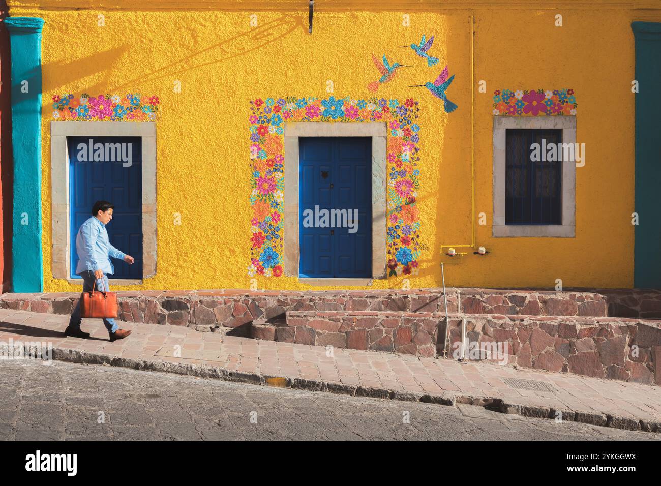 Guanajuato, Mexiko - 9. Februar 2024: Ein Mann geht an einer leuchtend gelben Wand vorbei, die mit einem bunten Blumengemälde und Kolibris geschmückt ist und Guanaj zeigt Stockfoto