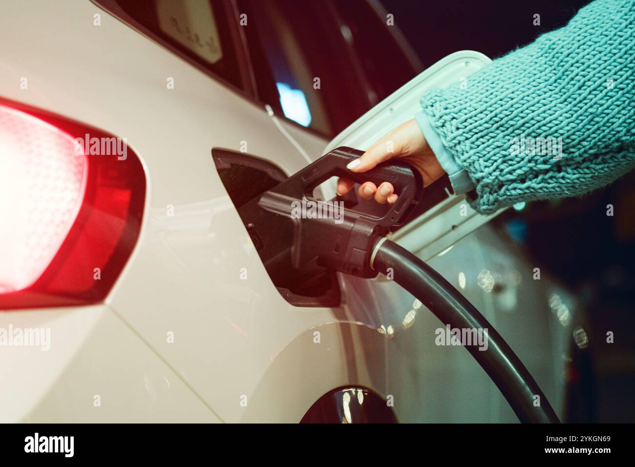 Eine Nahaufnahme einer Frauenhand, die ein Ladekabel an einem weißen Elektrofahrzeug an einer Ladestation anschließt und dabei saubere Energie und moderne Energie betont Stockfoto